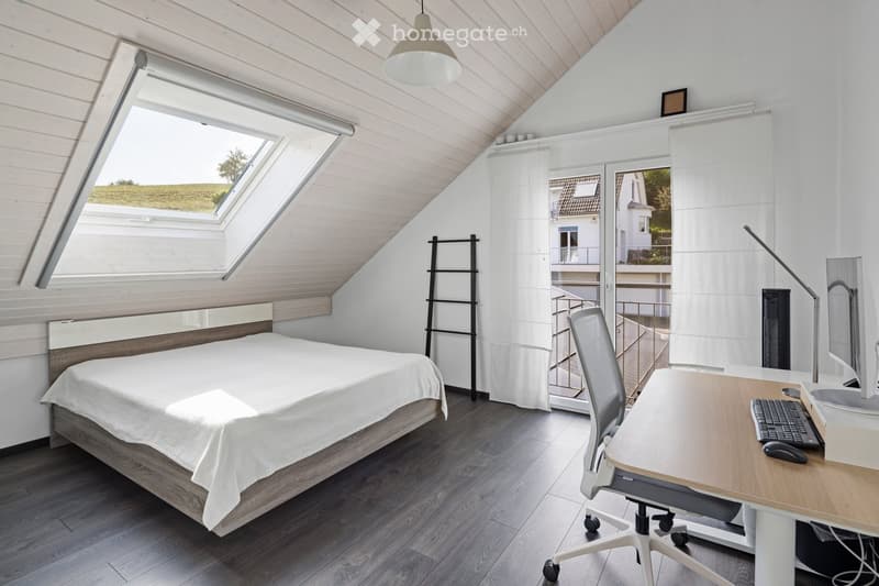 Bright bedroom with wooden slanted ceiling, large skylight windows, bed with white linens, desk with computer and chair, and a view of the outdoor area.