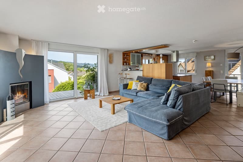 Modern living room with a grey sectional sofa, coffee table, fireplace, and large windows offering a scenic view. The space includes a dining area with a table and chairs, and an open kitchen with cabinets and a lamp.