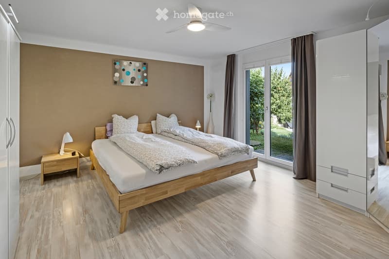Modern bedroom with a double bed, nightstand, lamp, wardrobe, and a large window with curtains. The room features wooden flooring and a contemporary aesthetic.