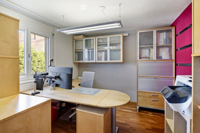 A modern office space with a wooden desk, ergonomic chair, mounted screen, and various cabinets and storage units.