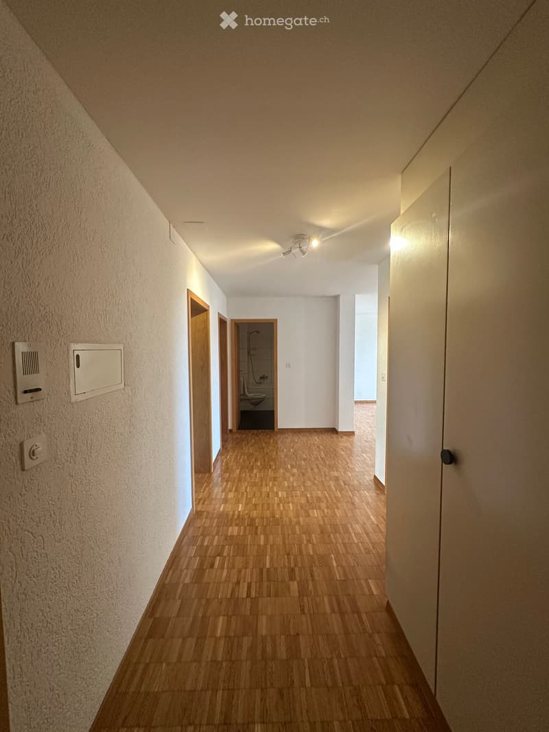 A narrow hallway with wooden flooring, white walls, and several doors leading to different rooms, including a visible bathroom with a toilet and sink.