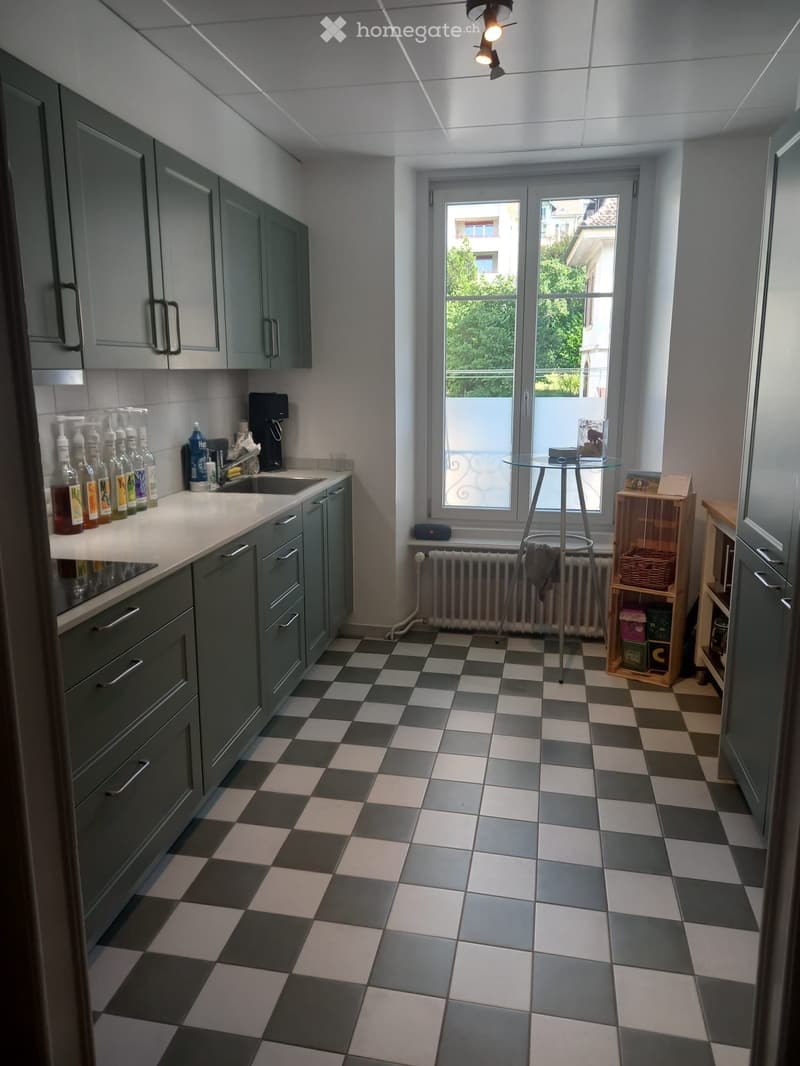 Modern kitchen with grey cabinets, checkered floor, and a large window with a view of greenery. Includes a sink, coffee maker, and a small round table with a stool.
