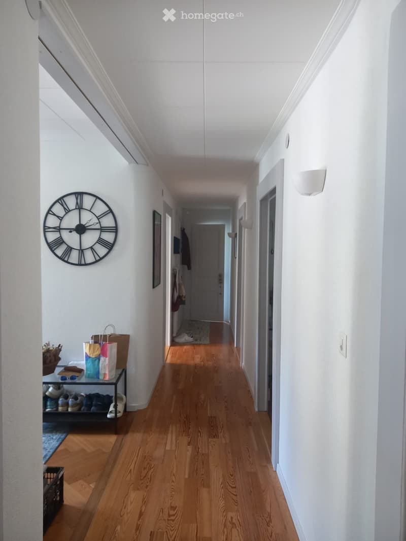 A well-lit hallway with wooden flooring, white walls, and a large wall-mounted clock. A small shoe rack and a shelf with bags are visible on the left side.