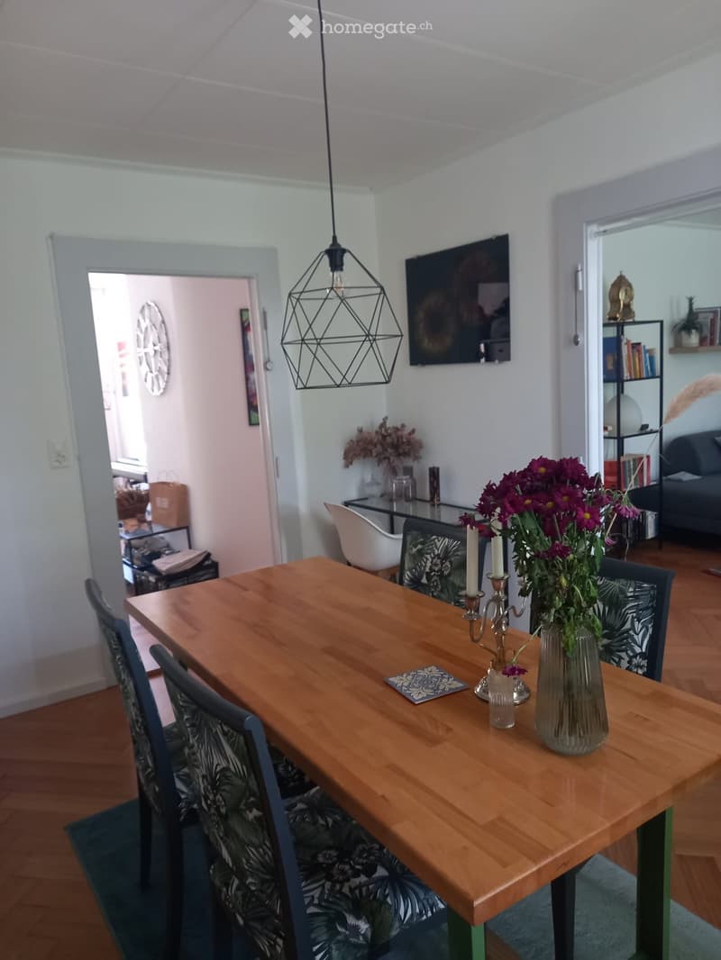 Dining area with a wooden table, chairs, vase with flowers, candle holder, and a TV in the background. The room has wooden flooring and white walls.