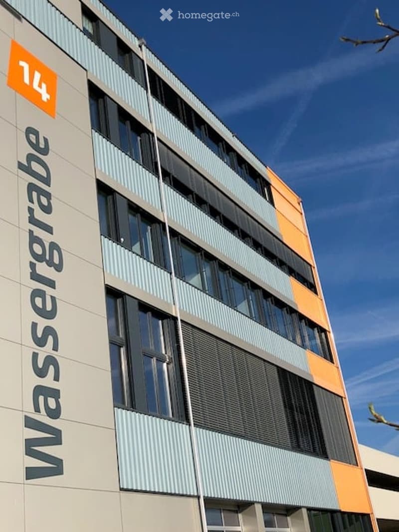 Modern multi-story building with a combination of light blue and orange panels, large windows, and signage reading "Wassergrabe" and the number "14".