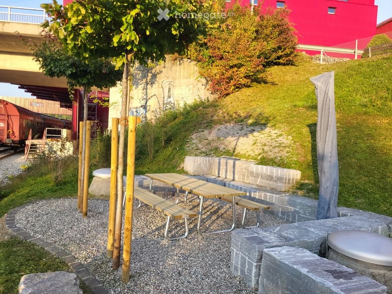 Outdoor seating area with wooden picnic tables and benches, surrounded by gravel and stone seating blocks, with a tree and a covered umbrella in the background.