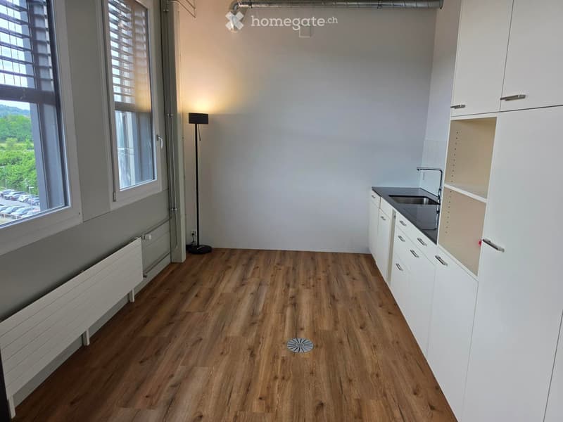 Empty room with wooden flooring, white cabinets, sink, and large windows with blinds. No furniture or appliances present.