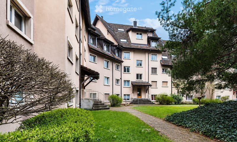A multi-story residential building with a beige facade, multiple windows, and a well-maintained garden area with green lawns and trimmed bushes.