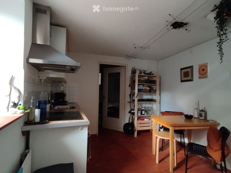 Modern kitchen with a stove, sink, and refrigerator. A small dining area features a table and chairs. Shelves hold various kitchen items, and framed pictures adorn the walls.