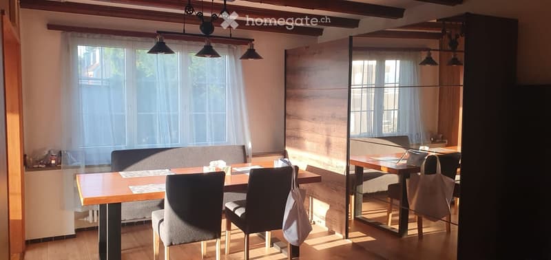 A modern, well-lit dining area with a wooden table, chairs, and a sofa, featuring exposed wooden beams and large windows with sheer curtains.