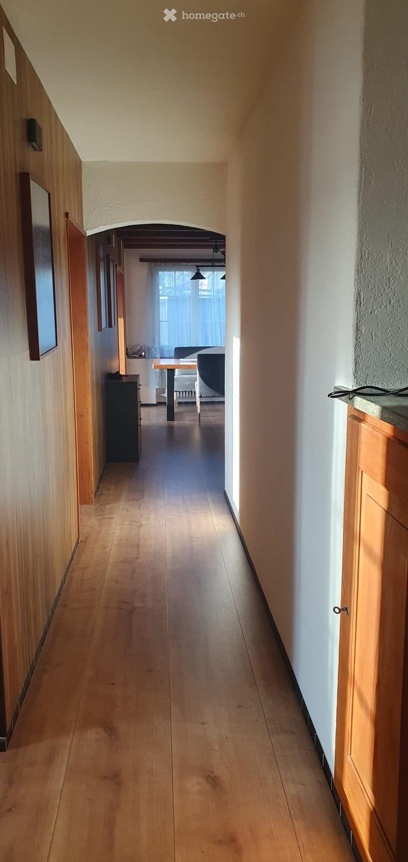 A long, narrow hallway with wooden flooring and white walls, featuring a wooden cabinet on the left and a small table and chair visible in the distance.