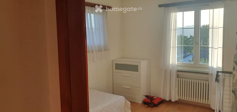 Bedroom with a bed, chest of drawers, radiator, and a drill on the floor. The room has wooden flooring, white walls, and two windows with white curtains.