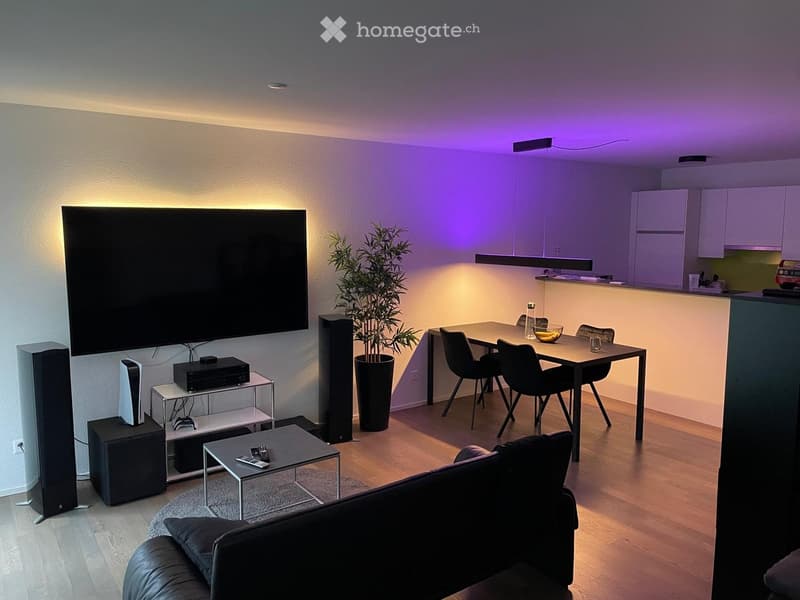 Modern living room with a large screen, sofa, coffee table, dining table with chairs, and a potted plant. The room features wooden flooring and ambient lighting.