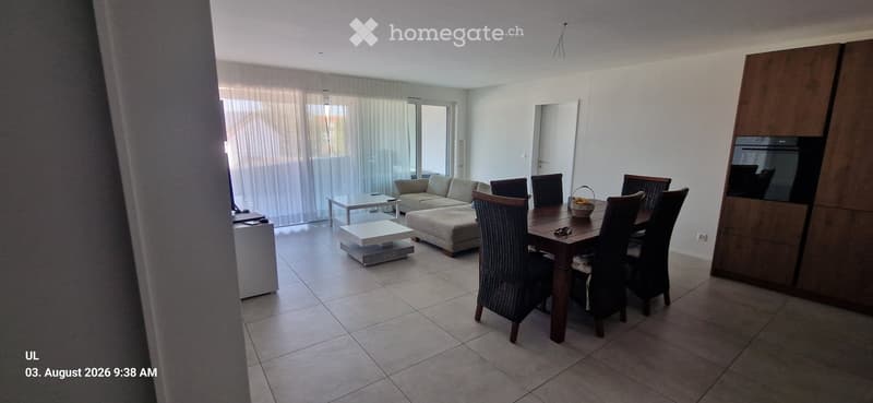 Bright living and dining area with a sofa, coffee table, ottoman, dining table, and chairs. Large windows with sheer curtains and a sliding glass door lead to a balcony.