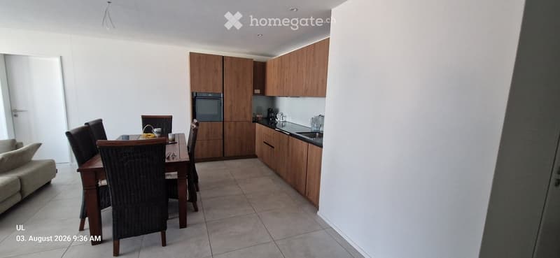 A modern, open-plan living and dining area with a wooden dining table, wicker chairs, a sofa, and a kitchen with wooden cabinets, an oven, and a sink.