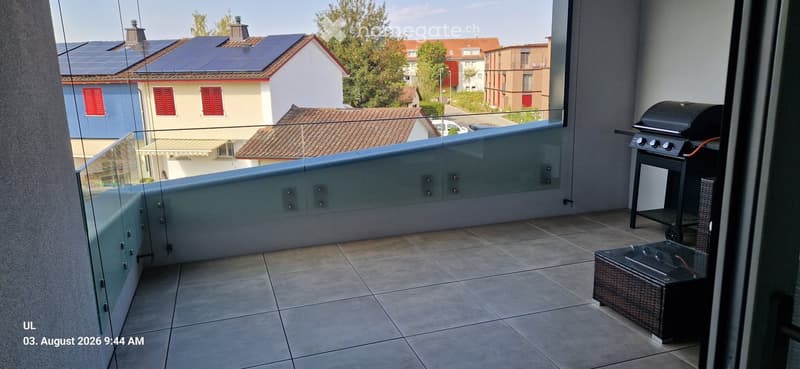 Balcony with tiled floor, glass railing, grill, and wicker chair.