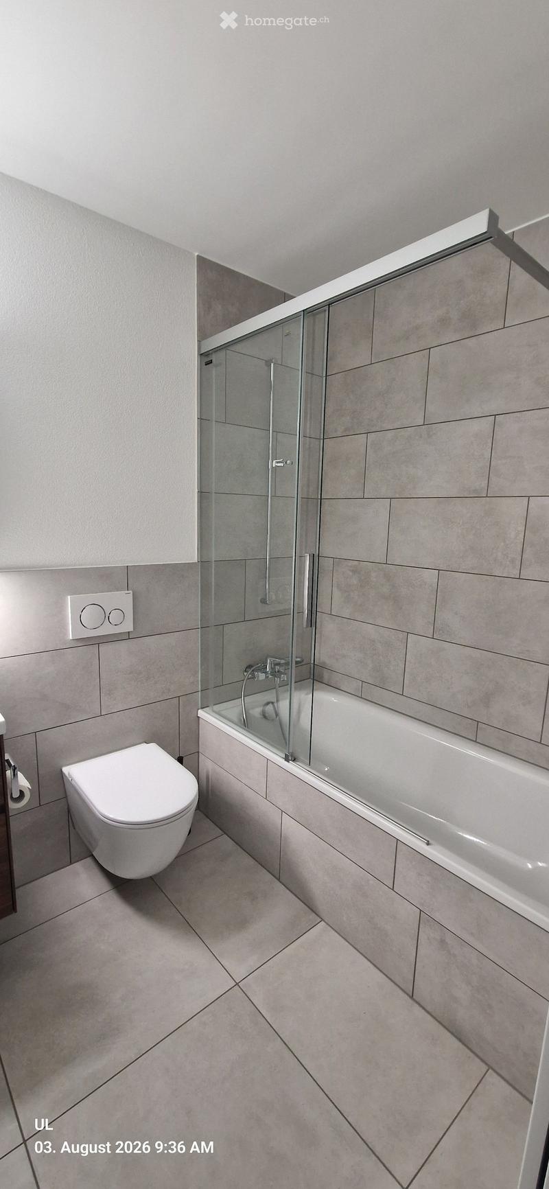 Modern bathroom with a white toilet, bathtub, and glass-enclosed shower area. Light gray tiles on walls and floor, sleek fixtures.