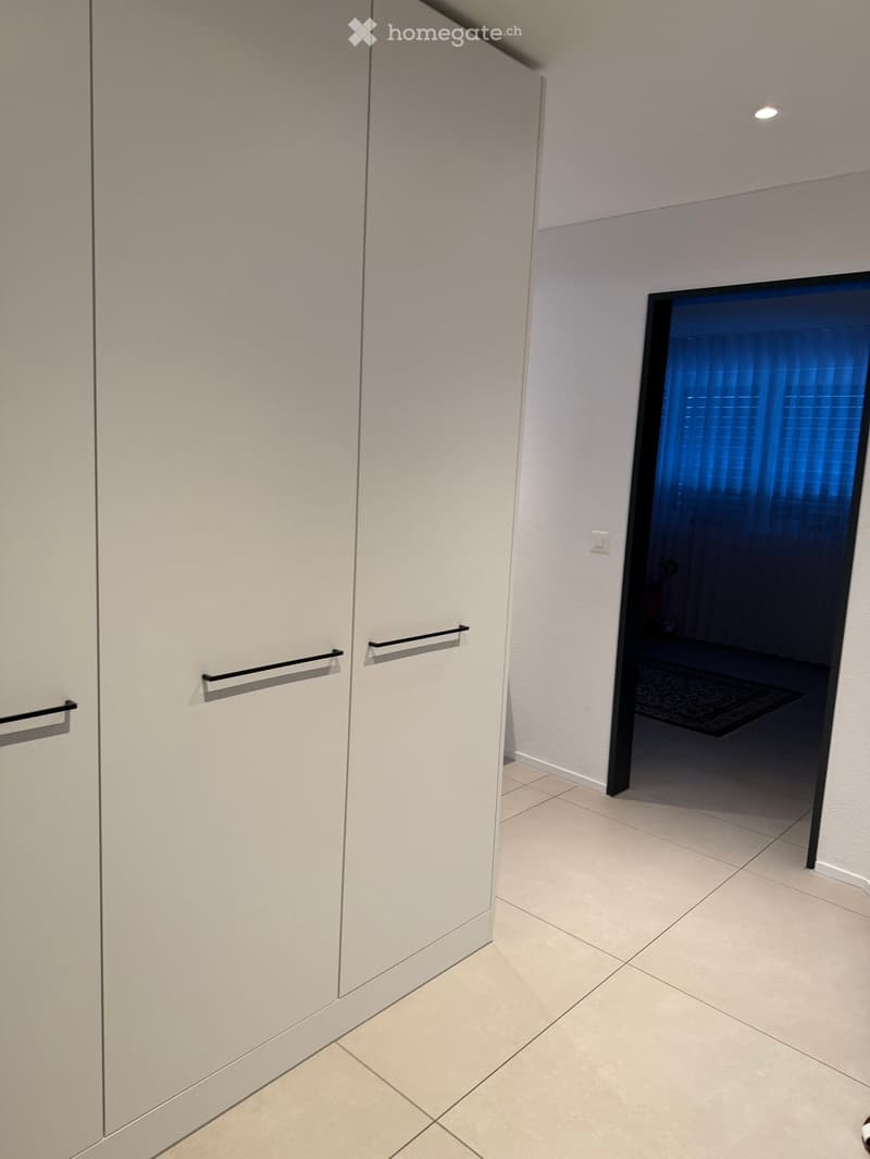 A modern hallway with light-colored tiled flooring, white walls, and a large mirrored closet with silver handles. A doorway leads to another room with a glimpse of a dark-colored rug and part of a piece of furniture.