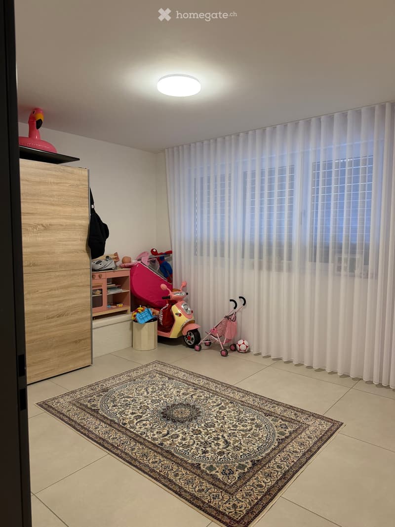 A well-lit room with a large window covered by white curtains, a patterned rug on the floor, a wooden wardrobe, a pink scooter, a baby walker, and various toys.