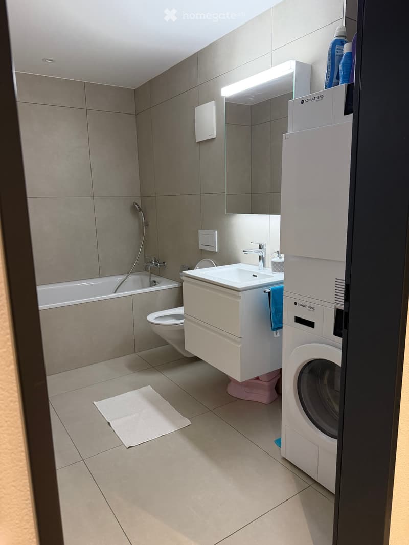 Modern bathroom with a bathtub, toilet, sink, mirror, and a washing machine. Features tiled walls and floor, a built-in vanity, and a showerhead.