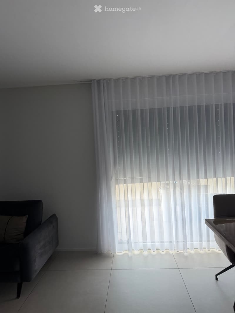 A modern, minimalistic living room with a grey sofa, a single chair, and a small table near the window. The room features white tiled flooring and large, sheer white curtains covering the window.