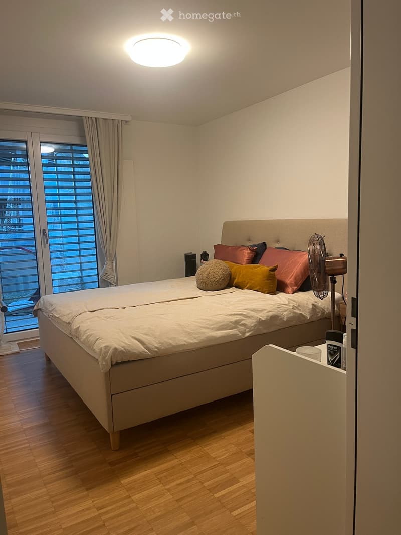 Modern bedroom with a large bed, colorful pillows, a standing fan, and a window with curtains. Wooden flooring and a built-in wardrobe are visible.