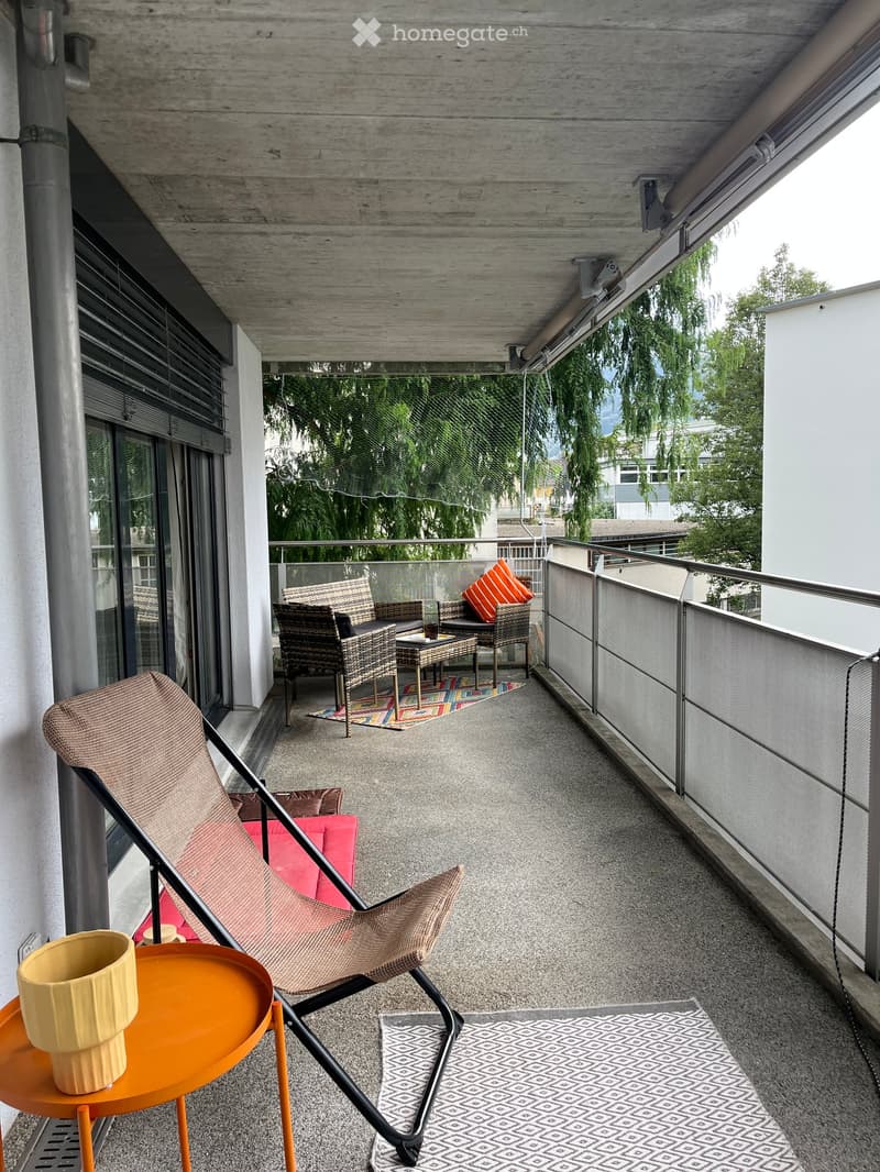 Balcony with modern furniture including a wicker chair, a small table, a sofa, and a cup on a small orange table. The balcony has a wooden ceiling and a metal railing with a view of greenery.