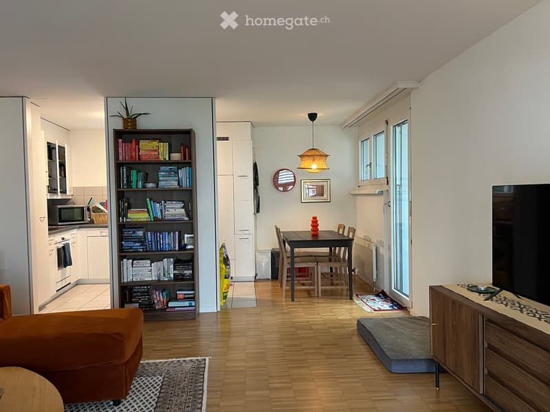 A modern, open-concept living and dining area with wooden flooring, a bookshelf filled with books, a sofa, a dining table with chairs, a kitchen area with appliances, and a TV stand with a screen.