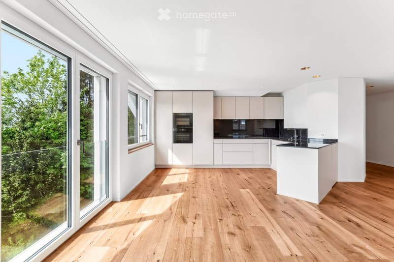 Modern kitchen with light-colored cabinets, wooden flooring, and large windows offering a view of greenery.