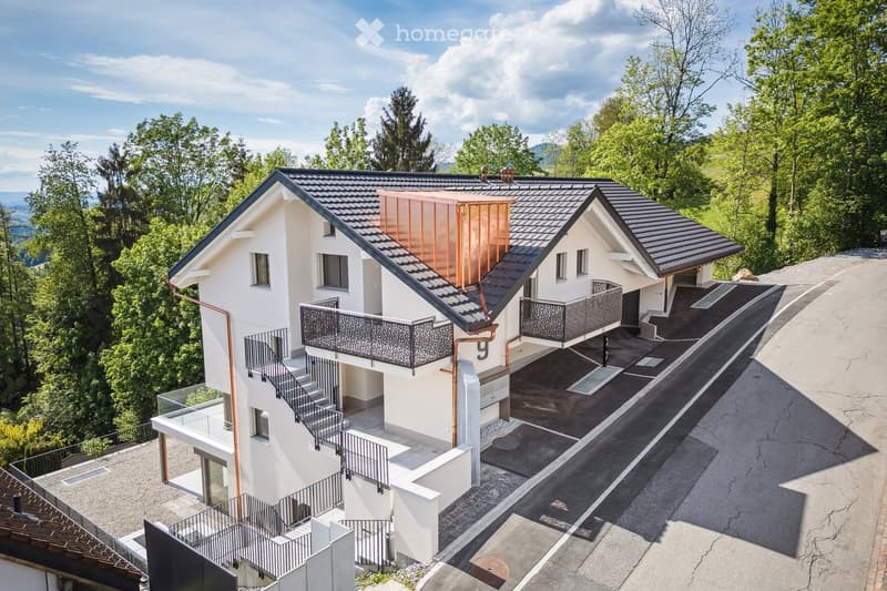 Modern two-story house with white exterior, dark grey roof, multiple balconies, and a raised ground floor. Surrounded by lush green trees and a scenic view.