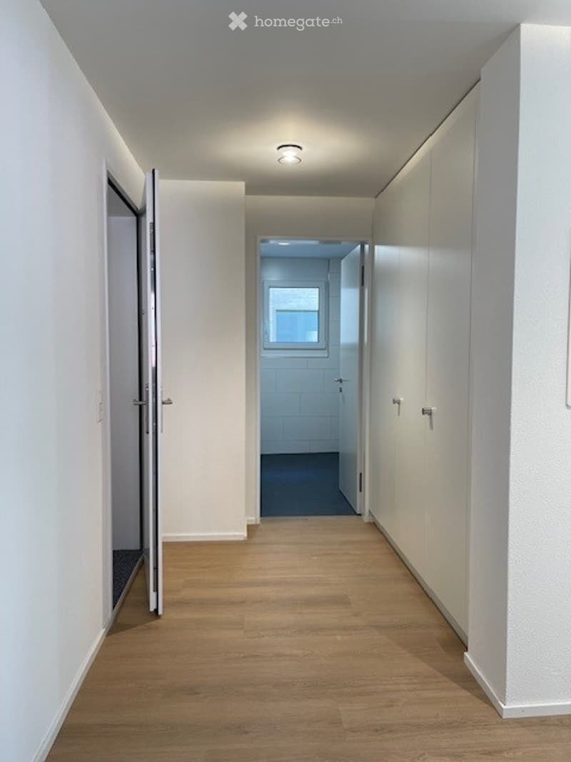 Clean, modern hallway with light wooden flooring, white walls, built-in storage cabinets, and recessed lighting. An open doorway leads to a room with a visible window.