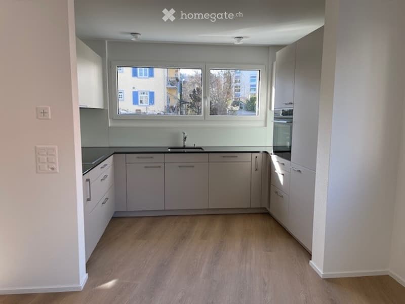 Modern kitchen with light grey cabinets, wooden flooring, and a large window overlooking a street.