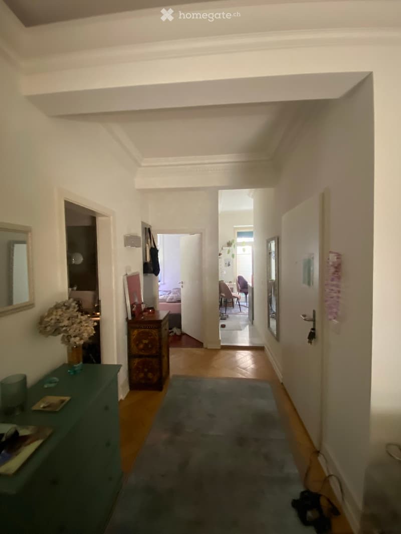 A well-lit hallway with wooden flooring, white walls, and several doors leading to different rooms. There is a small rug in the center, a green side table with a vase of flowers, and a decorative cabinet with ornate carvings.