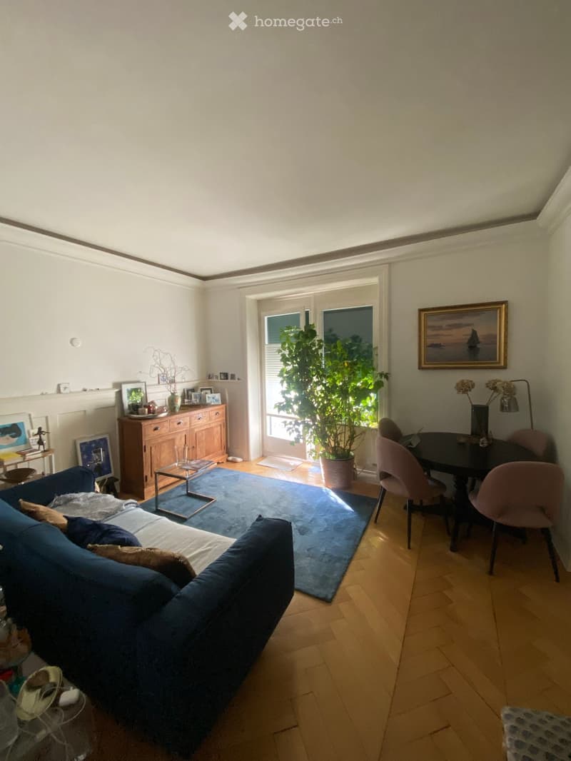 Bright living room with wooden flooring, blue sofa, coffee table, dining area with chairs, cabinet, large plant, and artwork on the walls.