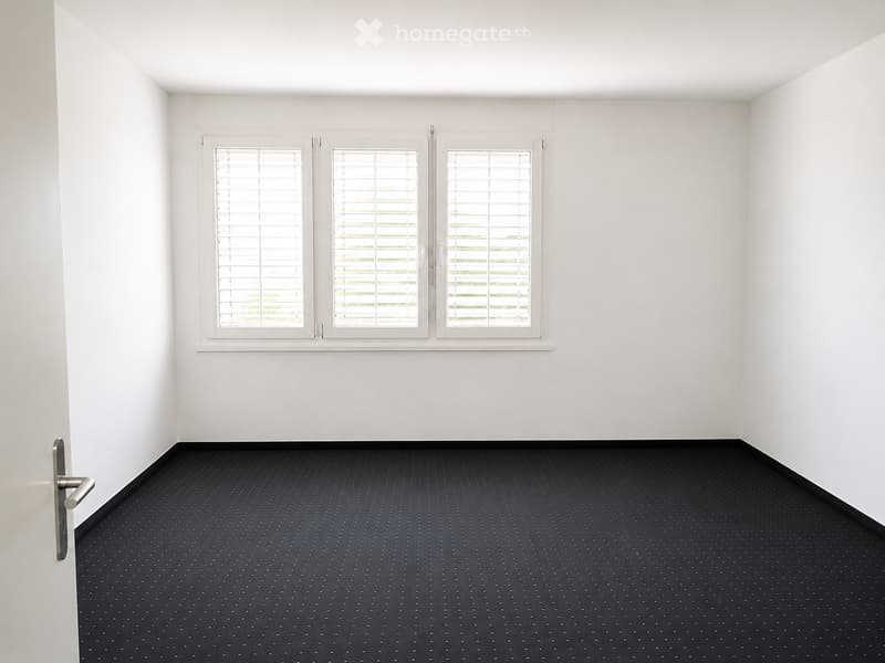 Empty room with white walls, dark patterned carpet, and large windows with blinds.