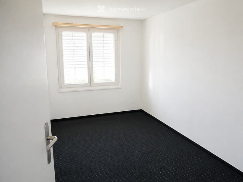Empty room with white walls, black carpet, and a double window with blinds. No furniture or equipment is present.