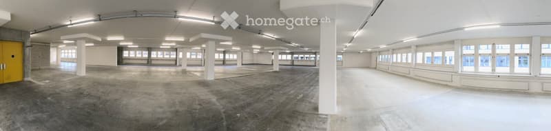Large, open, industrial-style space with exposed concrete floors, white walls, and large windows. Minimalist design with visible structural columns and a yellow door on the left side.
