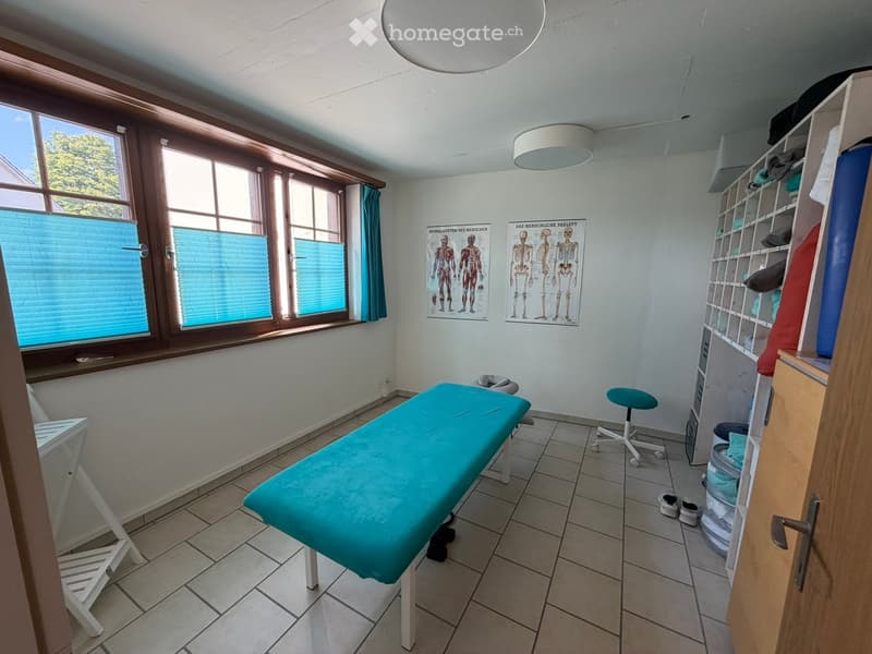 A clean, well-lit room with a treatment table, a small stool, shelves, and anatomical posters on the wall.