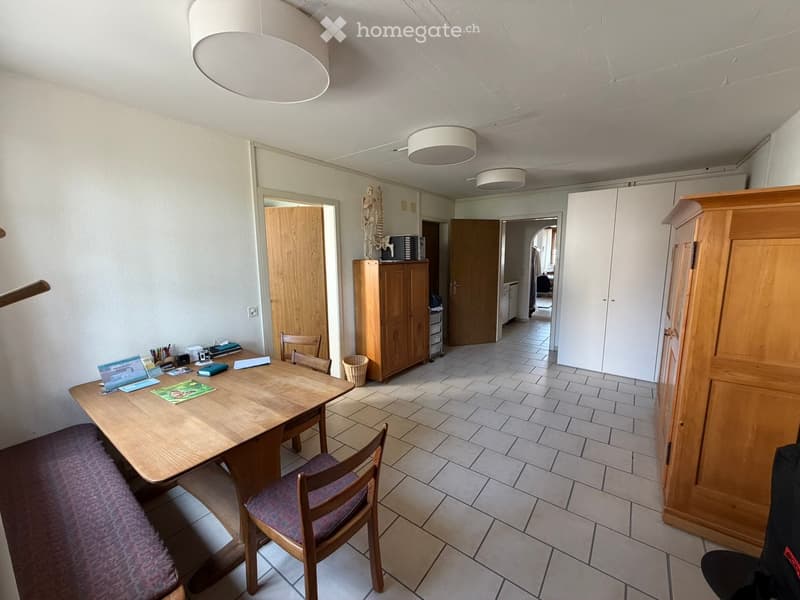 Bright, open dining area with a wooden table, chairs, and a cabinet. The space features tiled flooring and large windows, with additional furniture visible in the adjacent room.