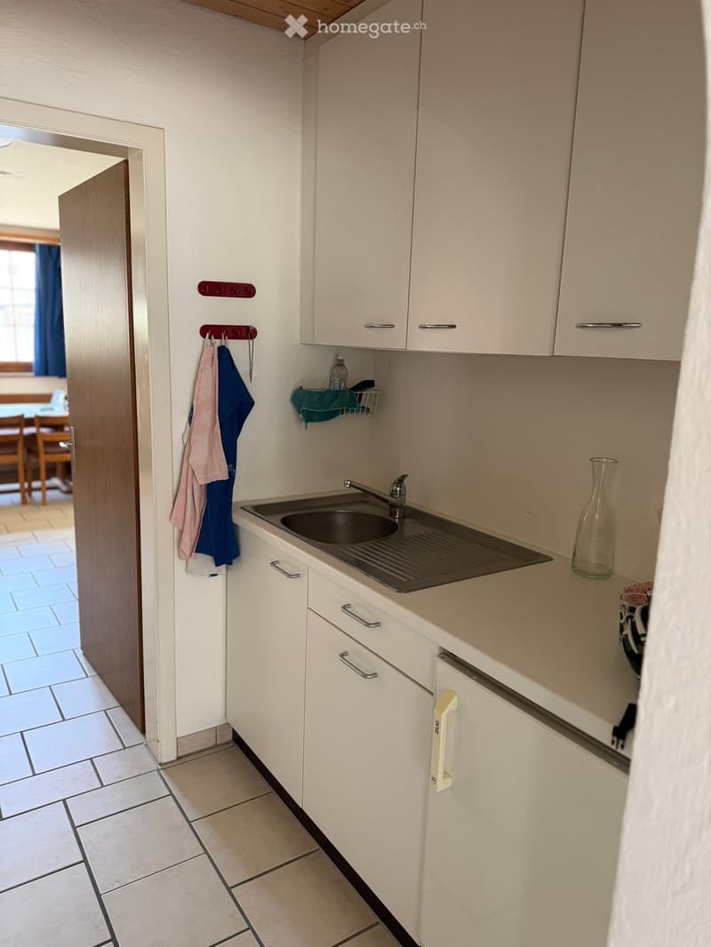Compact kitchen with white cabinets, a sink, and a countertop. A few items, including a bottle, cup, and vase, are placed on the counter. A doorway leads to another room with visible furniture.