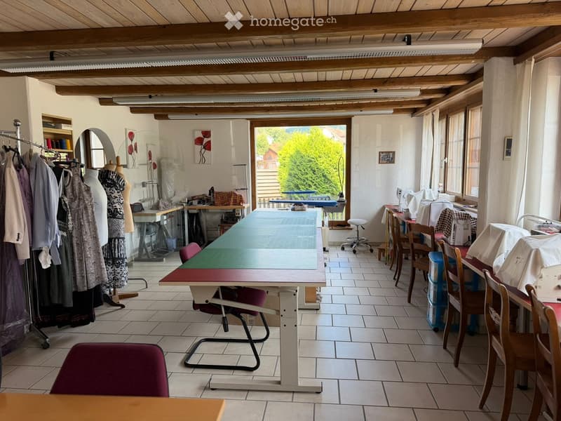 Brightly lit sewing or craft room with exposed wooden beams, large windows, tiled floor, and multiple work tables with chairs.