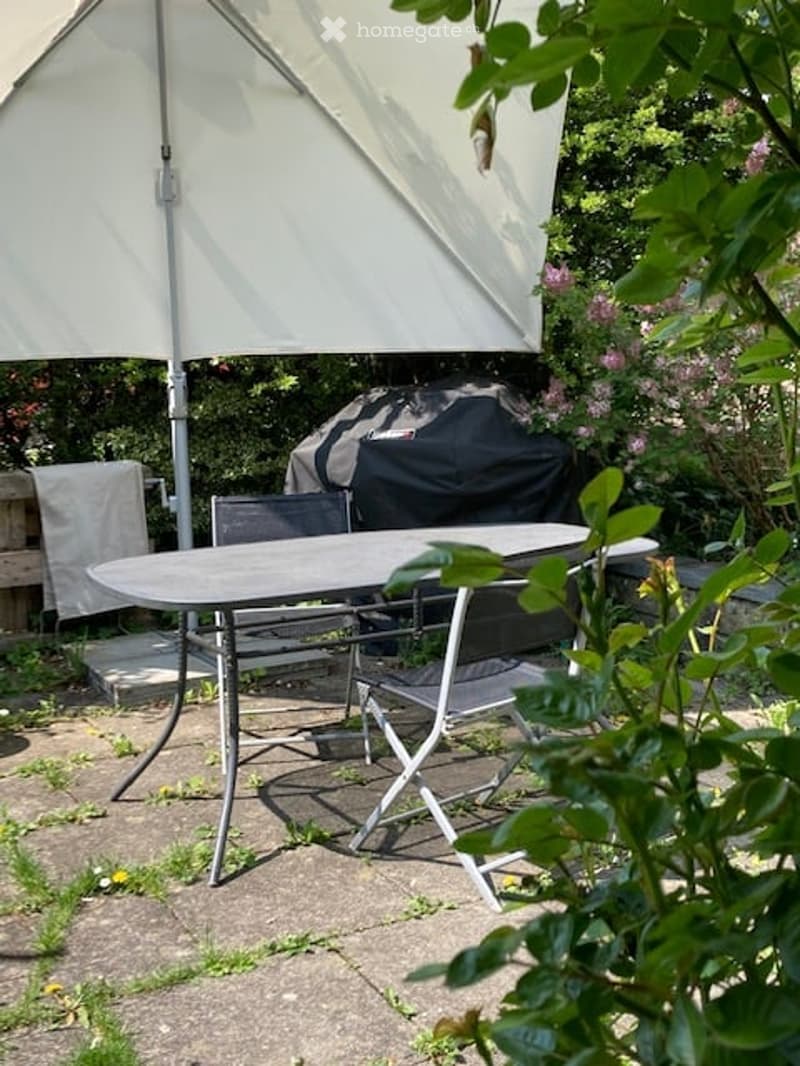 A patio area with a round table, four chairs, and an umbrella, surrounded by greenery and a covered grill.
