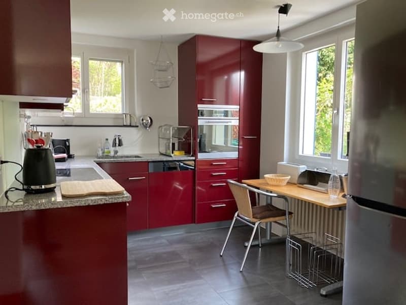 Modern kitchen with red cabinets, granite countertops, stainless steel appliances, and a small dining area with a table and chair.