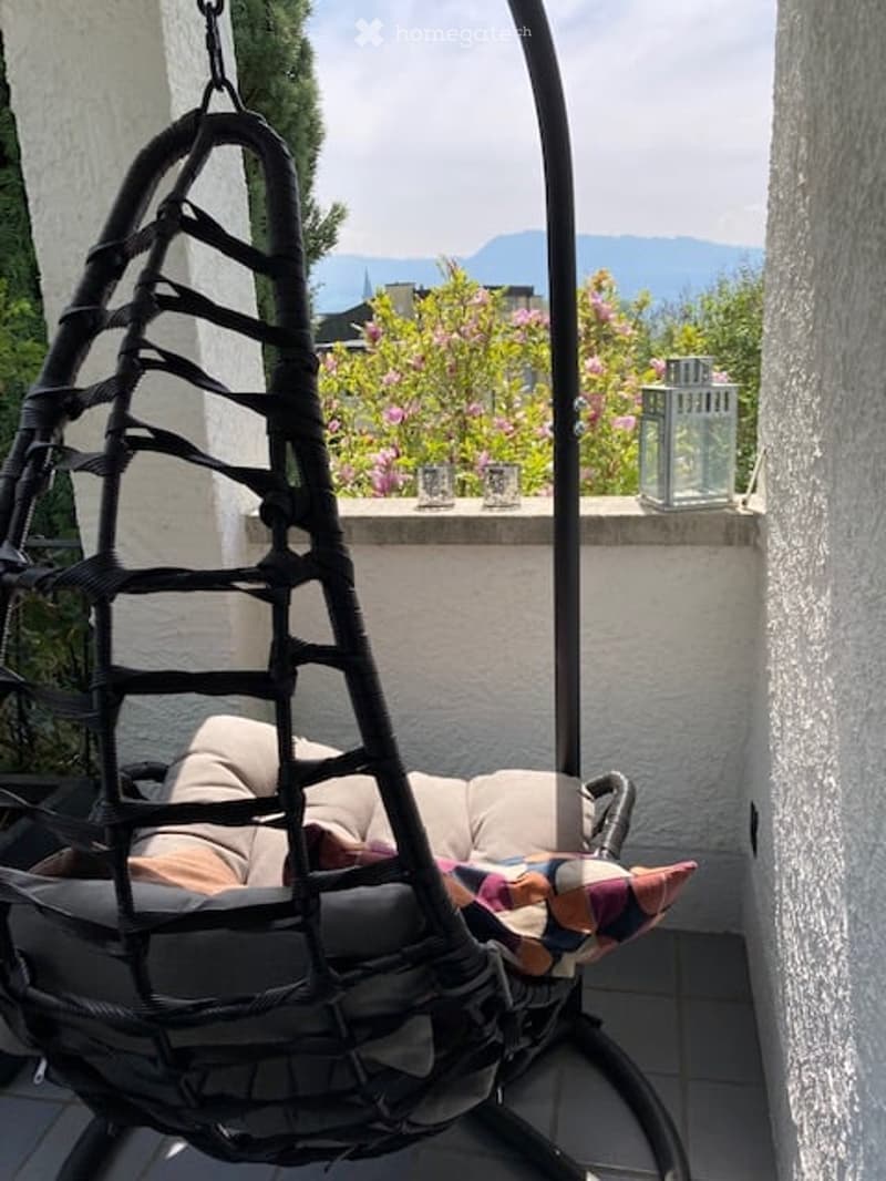 Balcony with a hanging egg chair, cushions, and a lantern, overlooking a garden with blooming flowers and distant mountains.