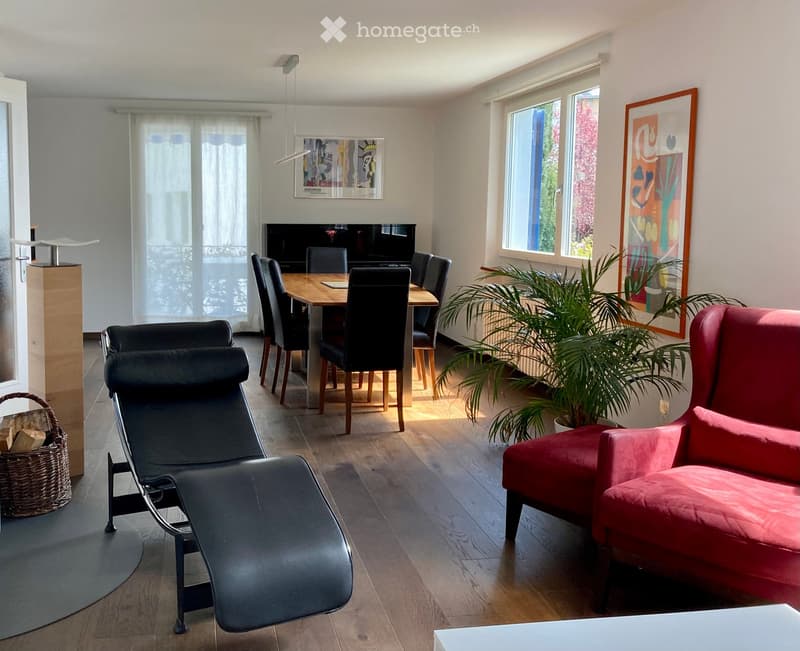 A modern living room with a red sofa, a black lounge chair, a wooden dining table with chairs, a large screen, and a potted plant near the window.