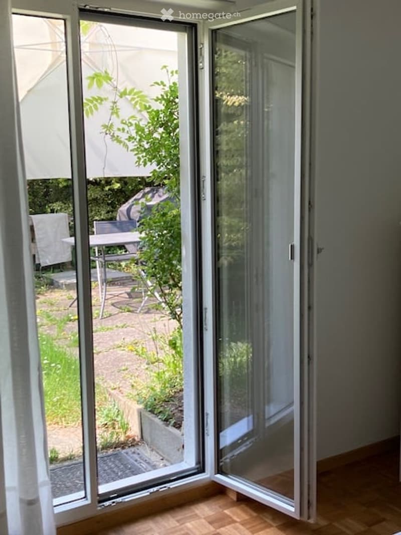 Open sliding glass doors leading to a small outdoor patio with a table and chairs, surrounded by greenery.