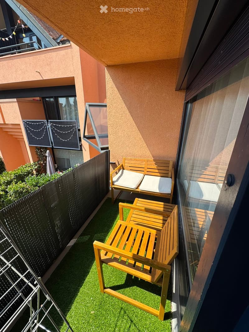 Balcony with artificial grass, wooden bench with white cushions, wooden chair, and a black privacy fence.