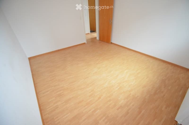 Empty room with light wooden flooring and white walls, featuring a wooden door leading to another room.