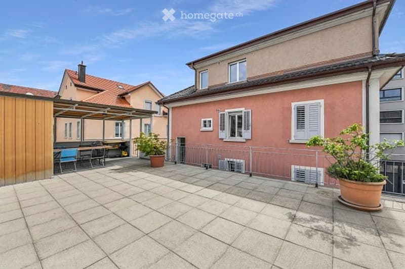 Spacious outdoor patio with tiled flooring, metal railing, potted plants, and a covered seating area with tables and chairs.