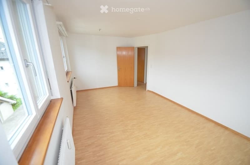 Empty room with light wooden flooring, white walls, large windows, and a wooden door leading to another room.